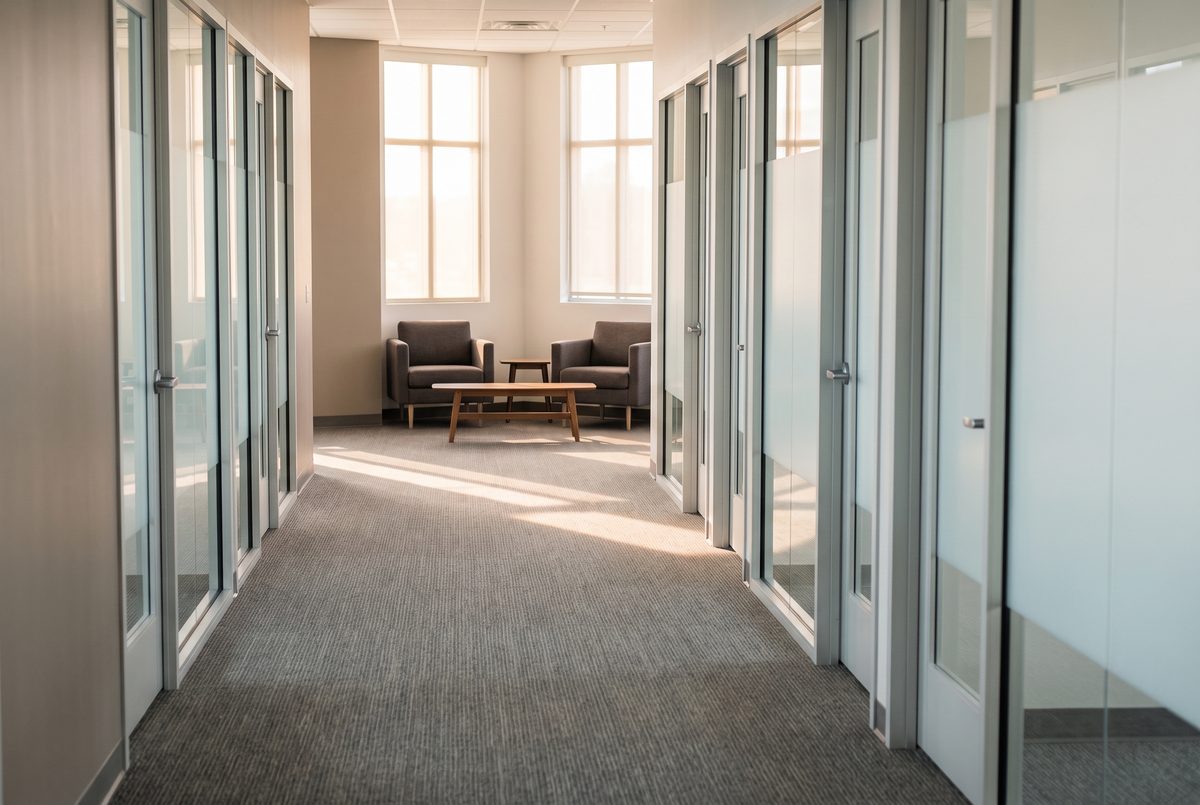 A bright shared office hallway with a carpeted corridor, glass suite doors and a lobby seating area in daylight